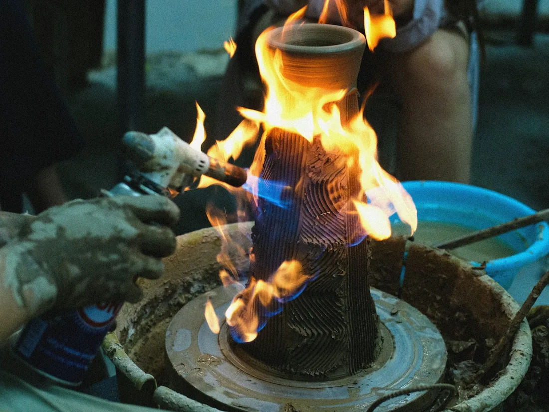 Pieza de barro rodeada de llamas durante una cocción de raku, con una mano enguantada sosteniendo un soplete cerca del cuello de la pieza